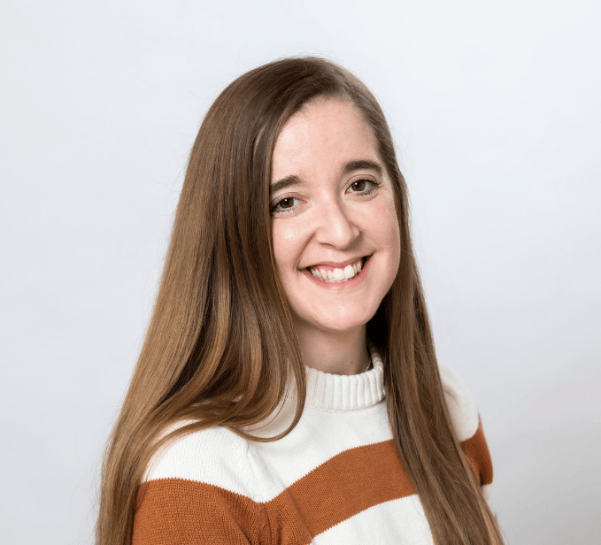 woman with medium brown long hair wearing a cream, rust and forest green large striped crewneck sweater smiles in a professional headshot