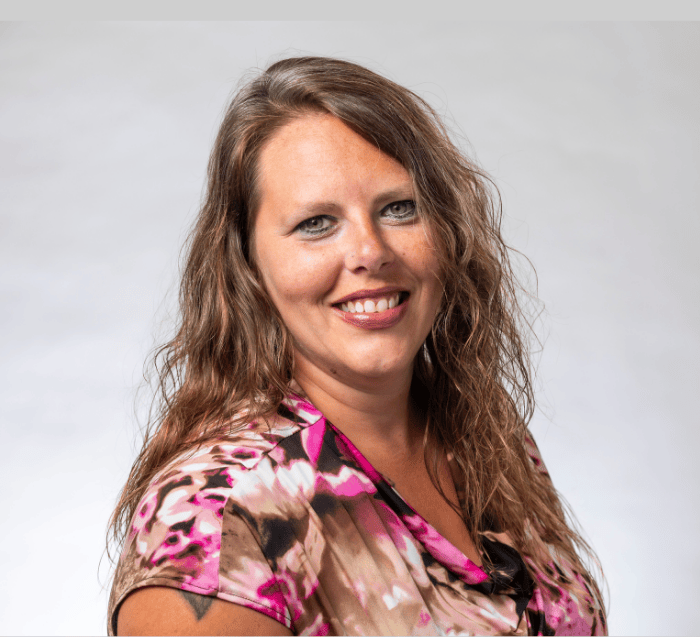 woman with wavy light brown hair wearing a pink, white, brown and black flowered v-neck blouse smiles in a professional headshot