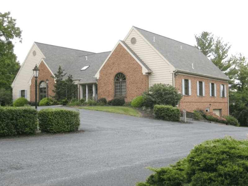 MFC Lancaster therapy office: light brown and tan building with a peaked rooftop with bushes and a blacktop driveway in front of it.