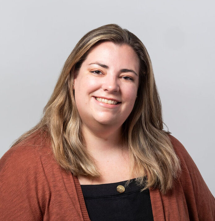 woman with brown hair and blond highlights wearing a black shirt, necklace and rust-colored cardigan smiling for a headshot