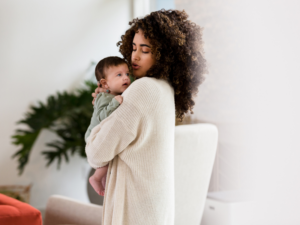 a mother with long curly black hair wearing a long white sweater holds a dark-haired baby boy in her arms in a white room with a house plant in the background
