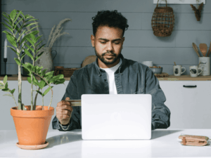 black man sitting in front of a laptop on a white table with a clay pot and green plant on it, looking at a virtual mental health counseling session.
