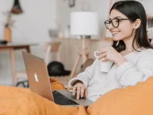 woman with black long hair and dark glasses holds a white coffeee cup and a laptop on her lap while leaning back on an orange couch in a living room doing a mental health therapy session
