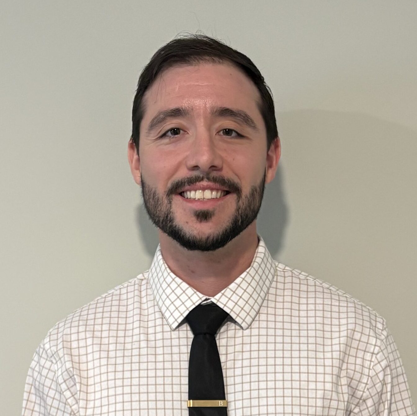 male mental health therapist with dark short hair, mustache and beard wearing a checked button-down dress shird and dark tie with a gold tie clip smiles for a professional headshot